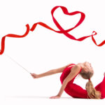 Isolated on white background Girl gymnast in a red overalls does exercise with a ribbon, the ribbon is twisted into a heart.