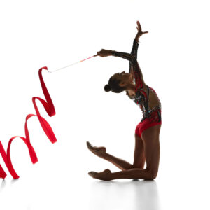 Artistic, talented teen girl, rhythmic gymnast performing with red ribbon, dancing against white studio background. Concept of sport, beauty and grace, competition, art, youth, hobby