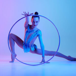 Beautiful teen girl, rhythmic gymnast, acrobat in stage costume dancing with hoop against gradient studio background in neon light. Concept of sport, beauty and grace, competition, art, youth, hobby